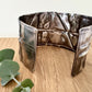 A wide, oxidised, foldformed sterling silver cuff bracelet. The cuff is highly textured and and is displayed on a wooden surface with a small sprig of eucalyptus leaves.