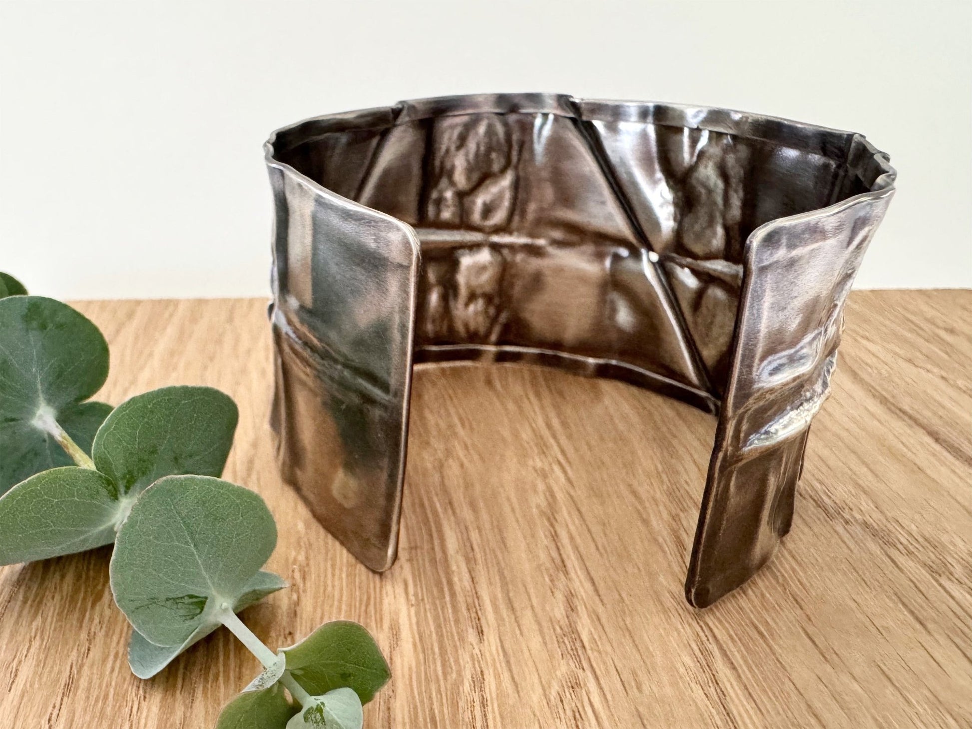 A wide, oxidised, foldformed sterling silver cuff bracelet. The cuff is highly textured and and is displayed on a wooden surface with a small sprig of eucalyptus leaves.