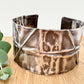 A wide, oxidised, foldformed sterling silver cuff bracelet. The cuff is highly textured and and is displayed on a wooden surface with a small sprig of eucalyptus leaves.