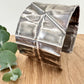 A wide, oxidised, foldformed sterling silver cuff bracelet. The cuff is highly textured and and is displayed on a wooden surface with a small sprig of eucalyptus leaves.