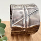 A wide, oxidised, foldformed sterling silver cuff bracelet. The cuff is highly textured and and is displayed on a wooden surface with a small sprig of eucalyptus leaves.