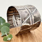 A wide, oxidised, foldformed sterling silver cuff bracelet. The cuff is highly textured and and is displayed on a wooden surface with a small sprig of eucalyptus leaves.