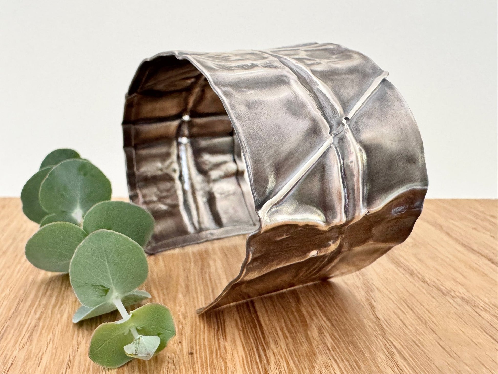 A wide, oxidised, foldformed sterling silver cuff bracelet. The cuff is highly textured and and is displayed on a wooden surface with a small sprig of eucalyptus leaves.