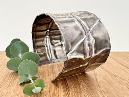 A wide, oxidised, foldformed sterling silver cuff bracelet. The cuff is highly textured and and is displayed on a wooden surface with a small sprig of eucalyptus leaves.