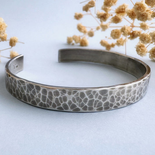 A heavy, hammered/beaten sterling silver cuff bracelet with a blackened finish is lying on a pale blue background. There are cream coloured dried flowers behind it.