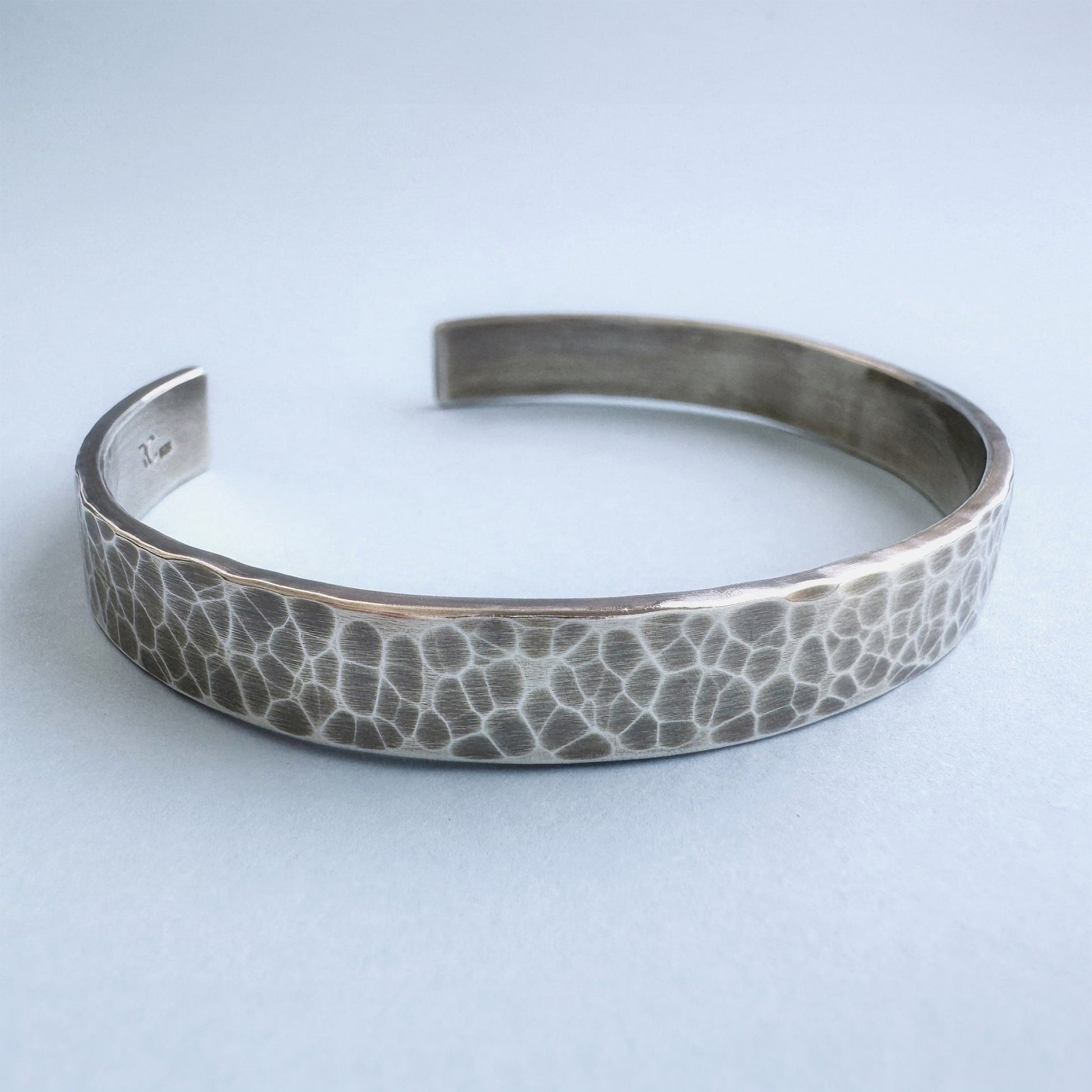 A heavy, hammered/beaten sterling silver cuff bracelet with a blackened finish is lying on a pale blue background.