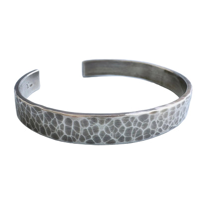 A heavy, hammered/beaten sterling silver cuff bracelet with a blackened finish is lying on a pale blue background.