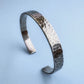 A heavy, hammered/beaten sterling silver cuff bracelet with a blackened finish is standing on a pale blue background. 