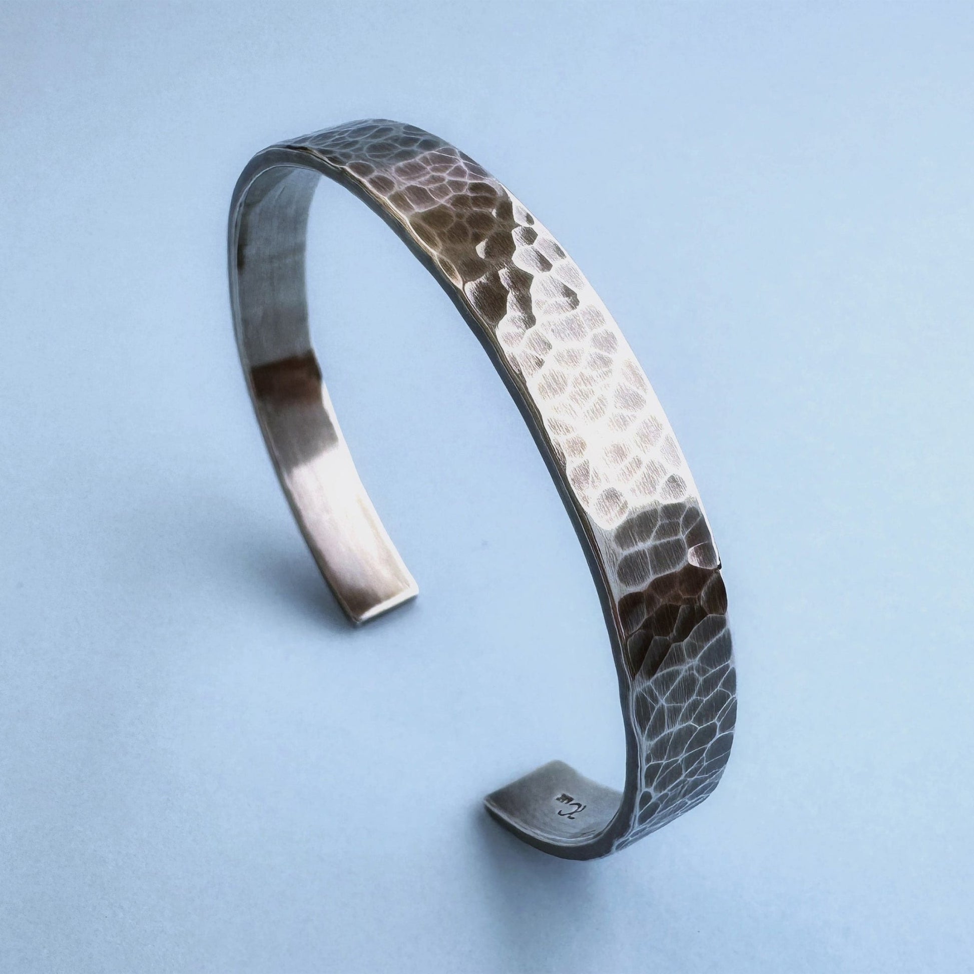 A heavy, hammered/beaten sterling silver cuff bracelet with a blackened finish is standing on a pale blue background. 