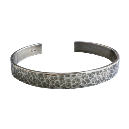 A heavy, hammered/beaten sterling silver cuff bracelet with a blackened finish is lying on a pale blue background.