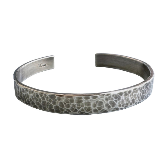 A heavy, hammered/beaten sterling silver cuff bracelet with a blackened finish is lying on a pale blue background.