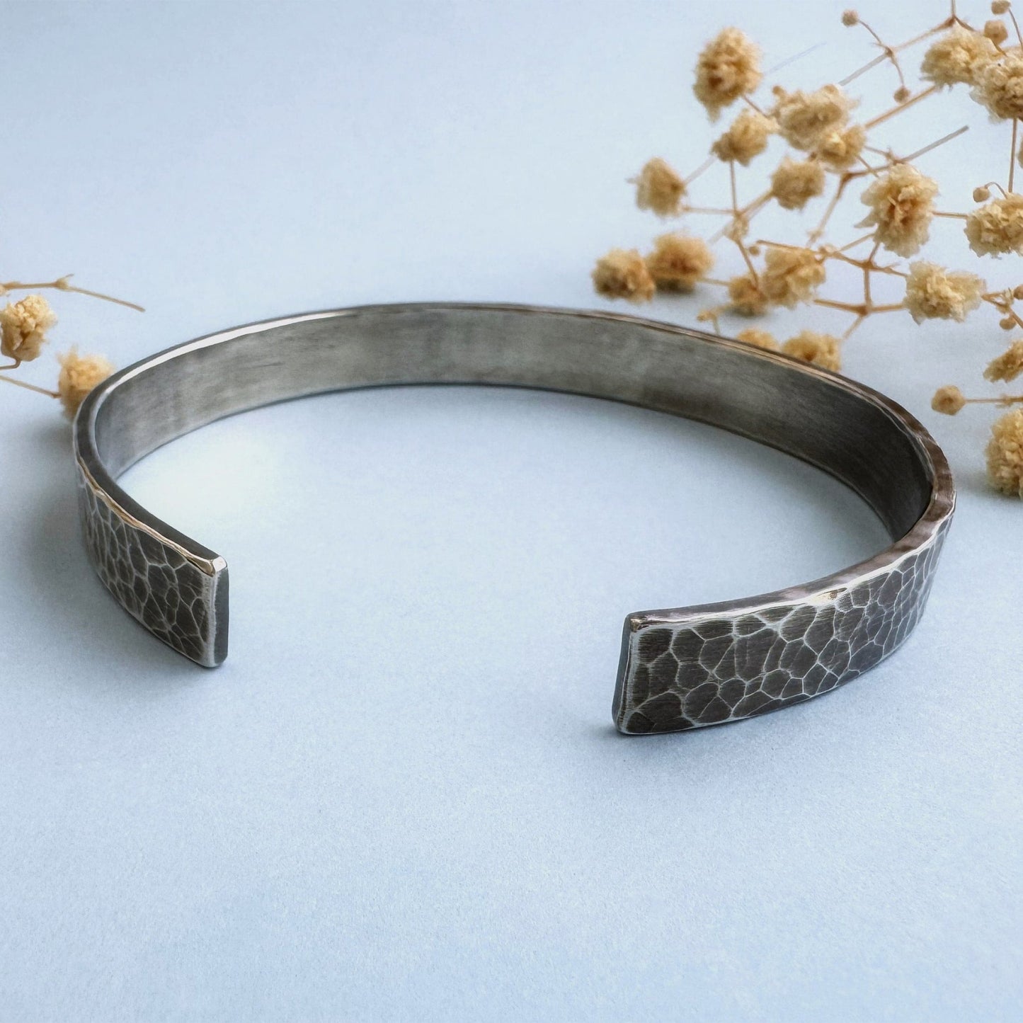 A heavy, hammered/beaten sterling silver cuff bracelet with a blackened finish is lying on a pale blue background. There are cream coloured dried flowers behind it.