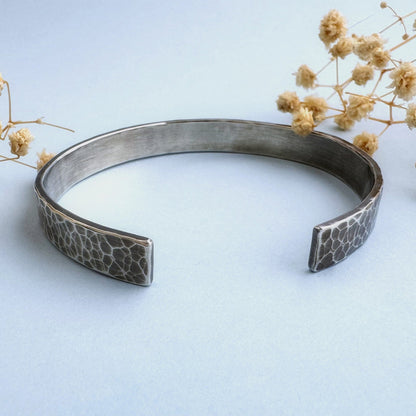 A heavy, hammered/beaten sterling silver cuff bracelet with a blackened finish is lying on a pale blue background. There are cream coloured dried flowers behind it.