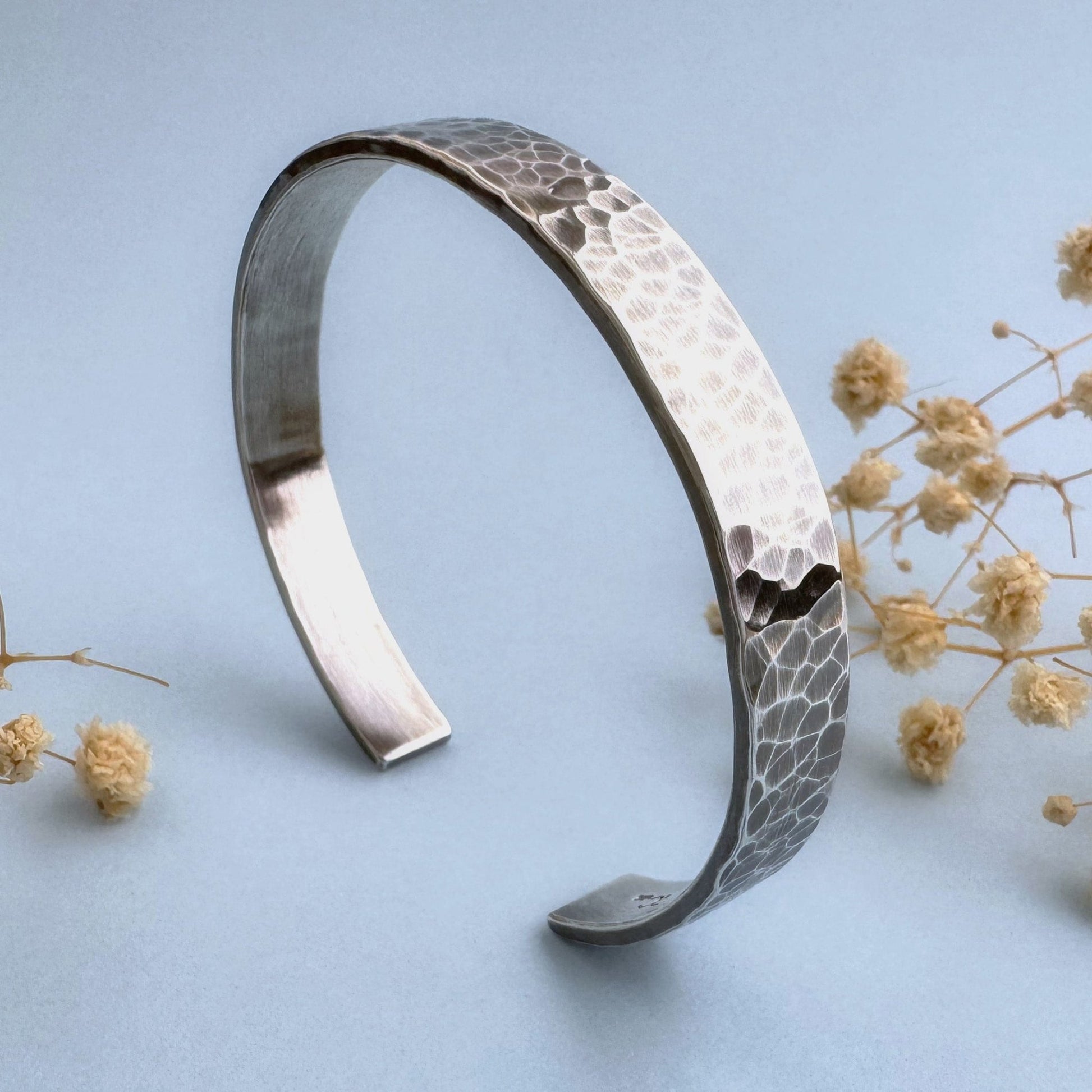 A heavy, hammered/beaten sterling silver cuff bracelet with a blackened finish is standing on a pale blue background. There are cream coloured dried flowers behind it.