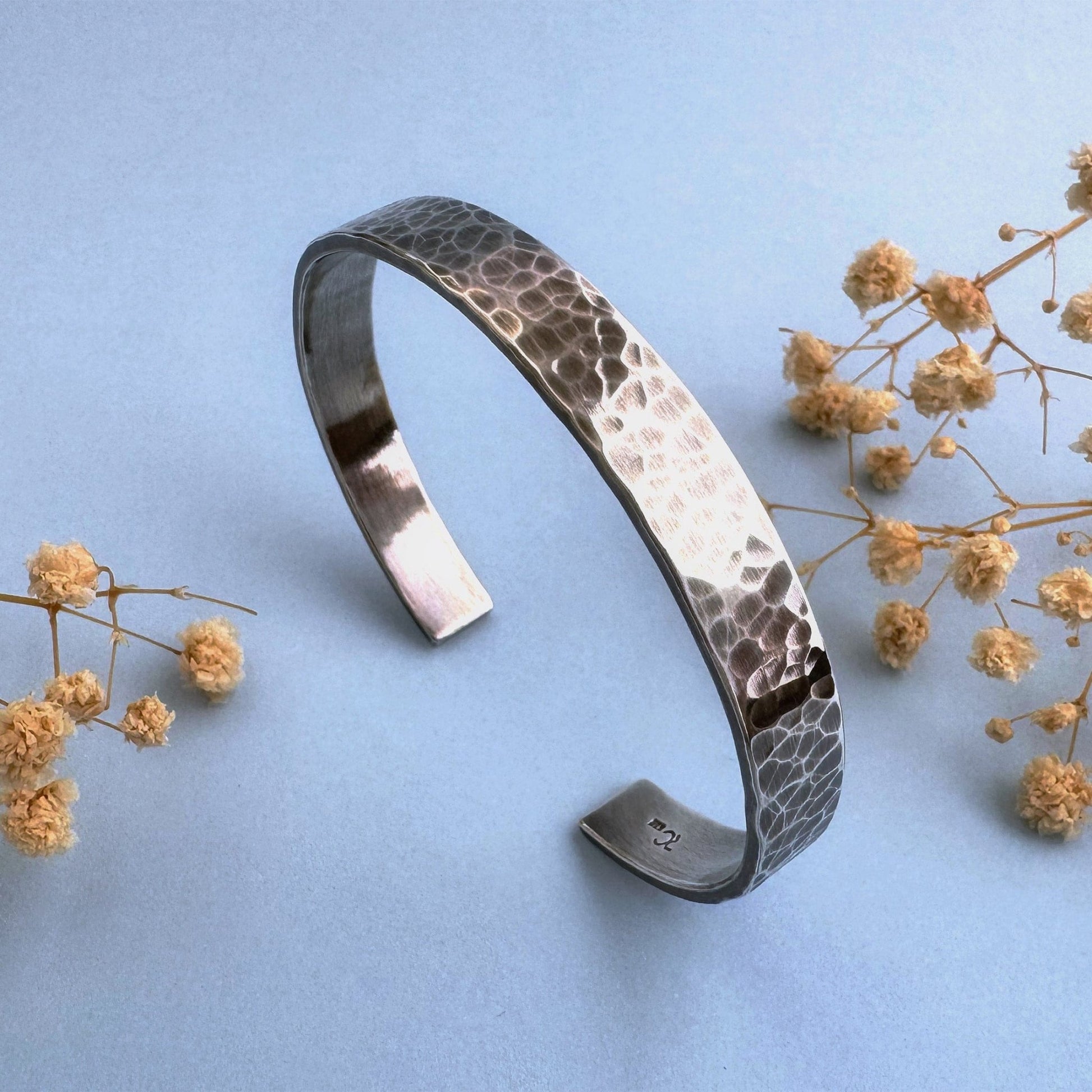 A heavy, hammered/beaten sterling silver cuff bracelet with a blackened finish is standing on a pale blue background. There are cream coloured dried flowers on either side of it.