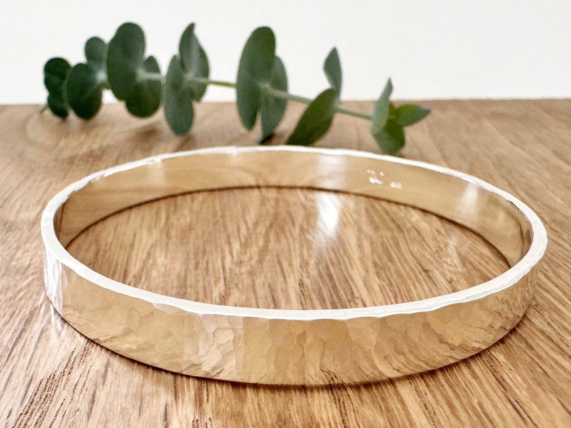 Hammered sterling silver bangle on a wooden surface with a small sprig of eucalyptus leaves