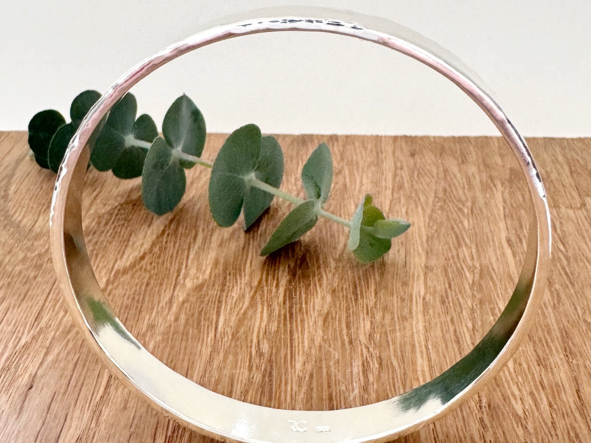 Hammered sterling silver bangle on a wooden surface with a small sprig of eucalyptus leaves