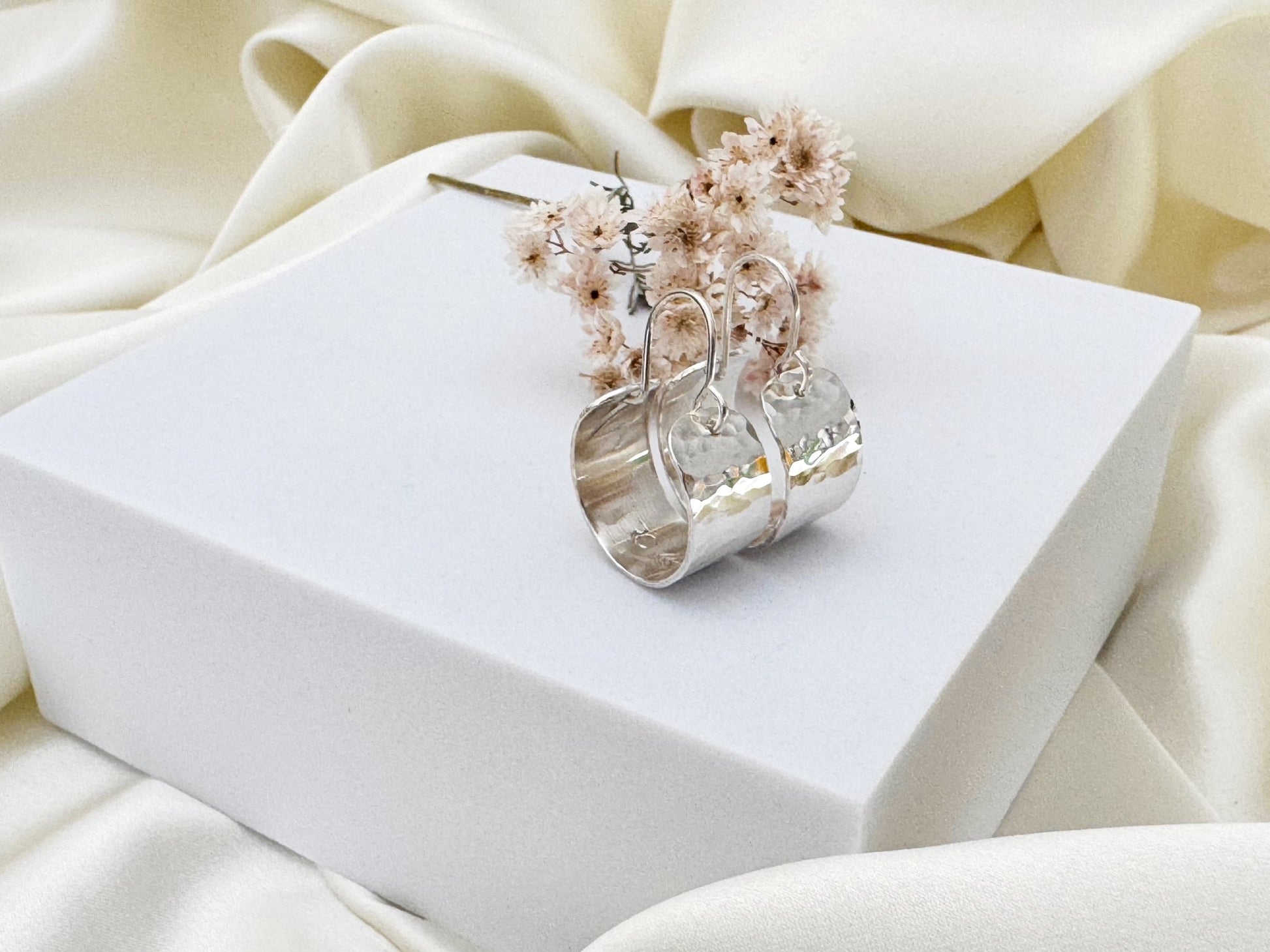 Silver hoop earrings with hammered texture on a white box against a beige fabric background