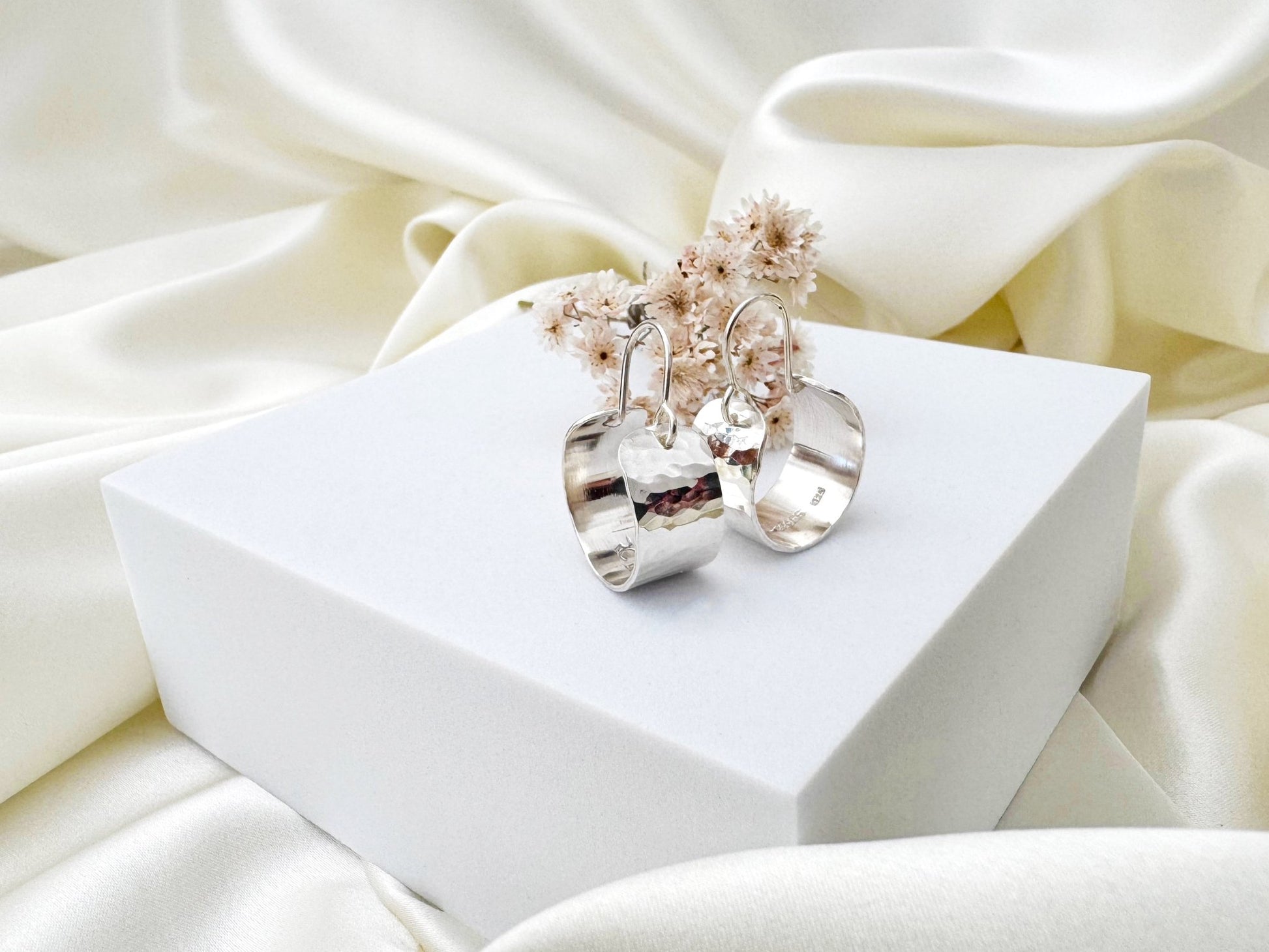 Silver hoop earrings with a hammered texture on a white box against a soft white fabric background