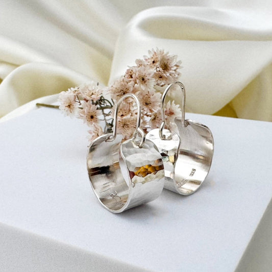 Silver hoop earrings with hammered texture on a white surface in front of a cream satin background