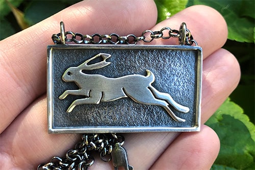 A handmade rectangular sterling silver pendant with a raised hare running from right to left, held in front of green leaves | Rebecca Cordingley Jewellery