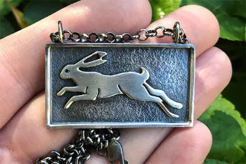 A handmade rectangular sterling silver pendant with a raised hare running from right to left, held in front of green leaves | Rebecca Cordingley Jewellery