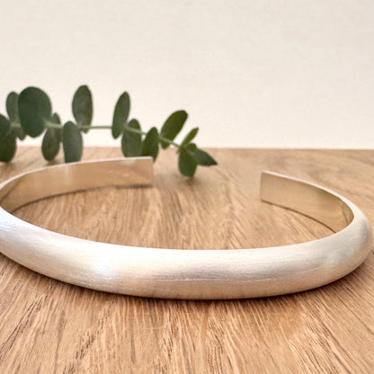 Heavy half-round sterling silver cuff bracelet on a wooden surface with a small sprig of eucalyptus leaves.
