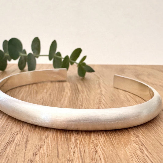 Heavy half-round sterling silver cuff bracelet on a wooden surface with a small sprig of eucalyptus leaves.