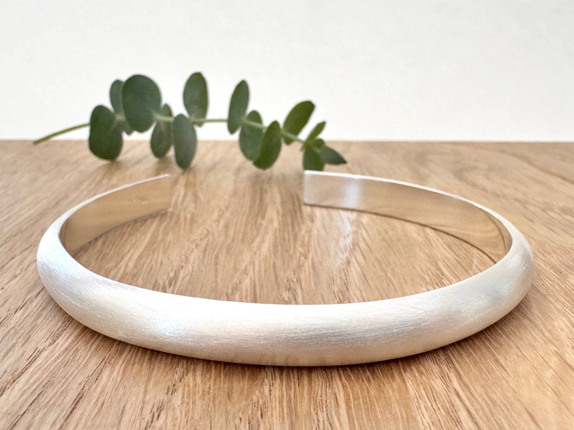 Heavy half-round sterling silver cuff bracelet on a wooden surface with a small sprig of eucalyptus leaves.