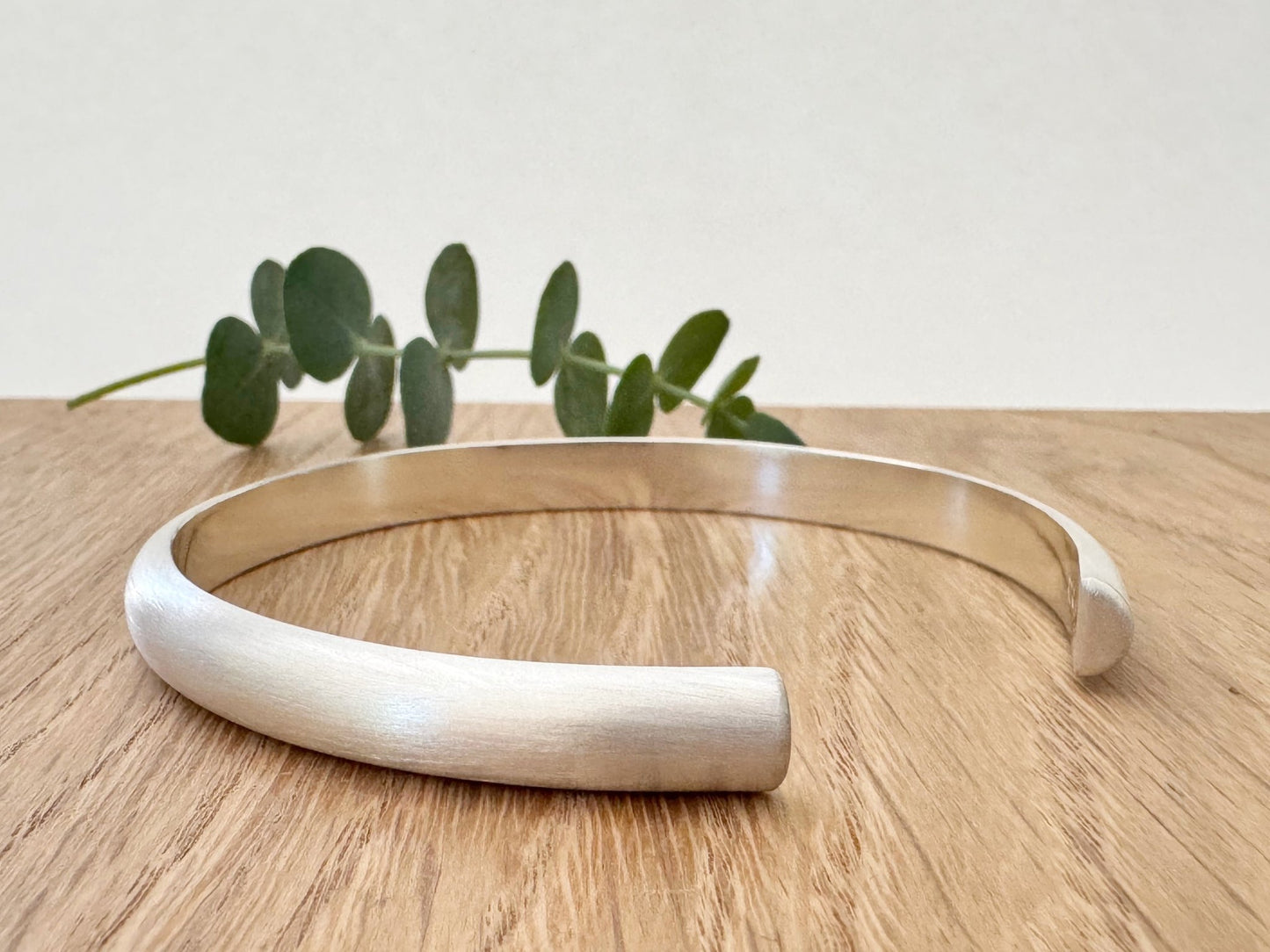 Heavy half-round sterling silver cuff bracelet on a wooden surface with a small sprig of eucalyptus leaves.