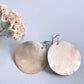 
A pair of large, hammered sterling silver disc earrings rests on a light blue background next to a small sprig of dried pink and white flowers.