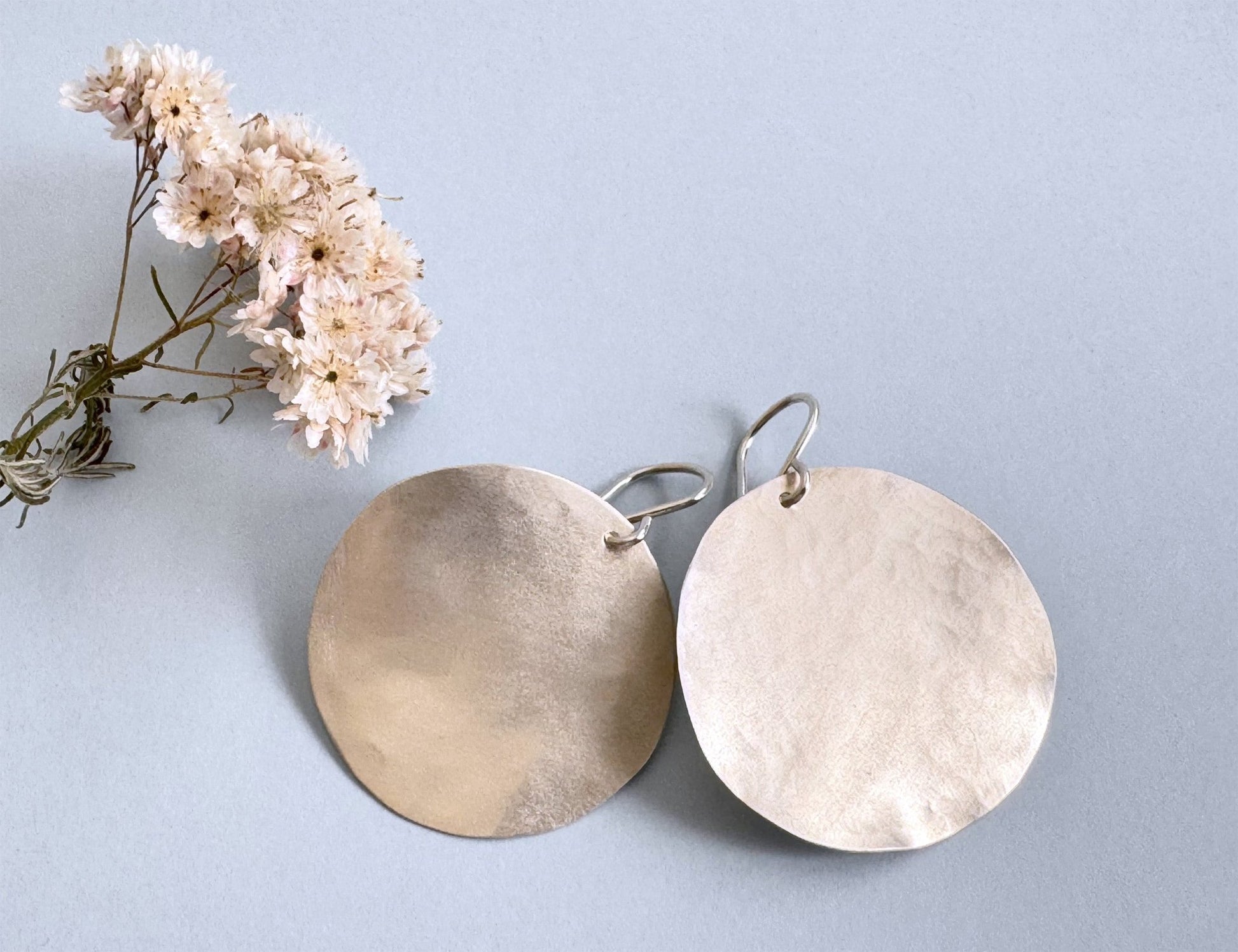 
A pair of large, hammered sterling silver disc earrings rests on a light blue background next to a small sprig of dried pink and white flowers.