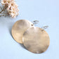 
A pair of large, hammered sterling silver disc earrings rests on a light blue background next to a small sprig of dried pink and white flowers.