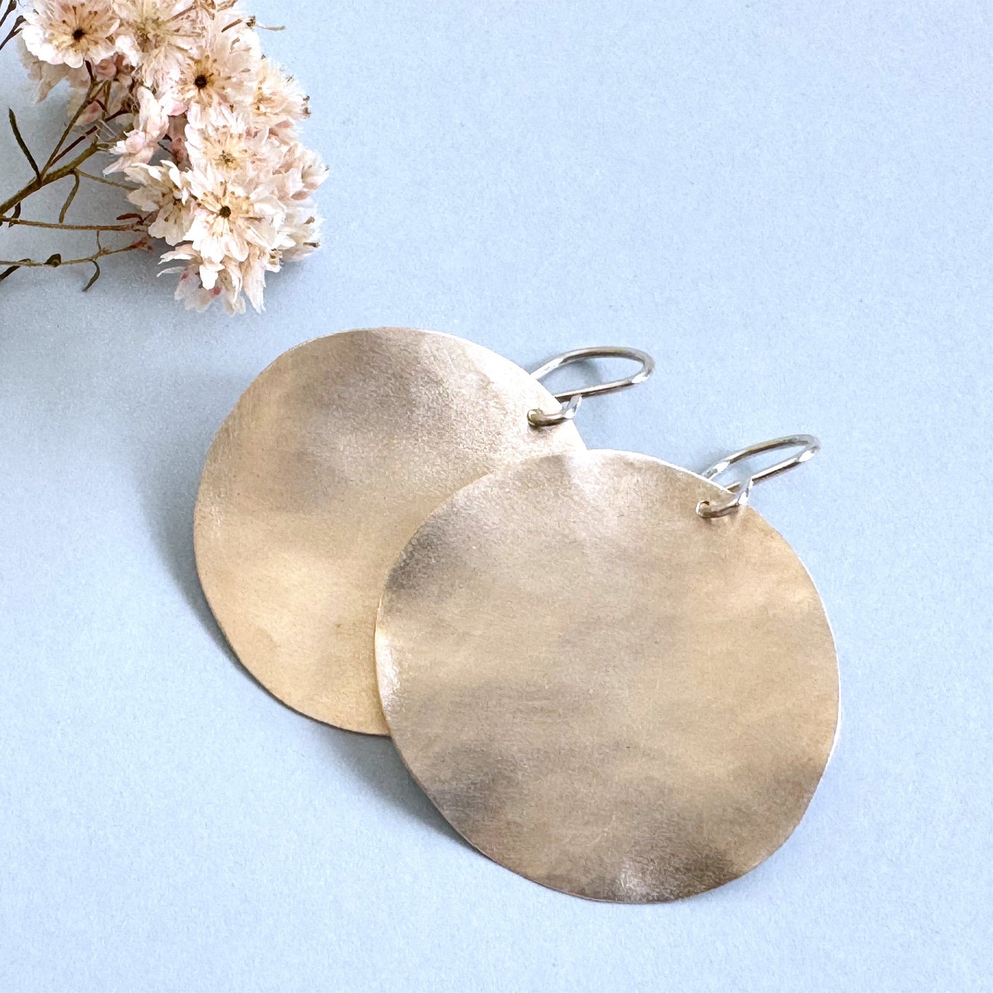 
A pair of large, hammered sterling silver disc earrings rests on a light blue background next to a small sprig of dried pink and white flowers.