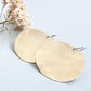 
A pair of large, hammered sterling silver disc earrings rests on a light blue background next to a small sprig of dried pink and white flowers.