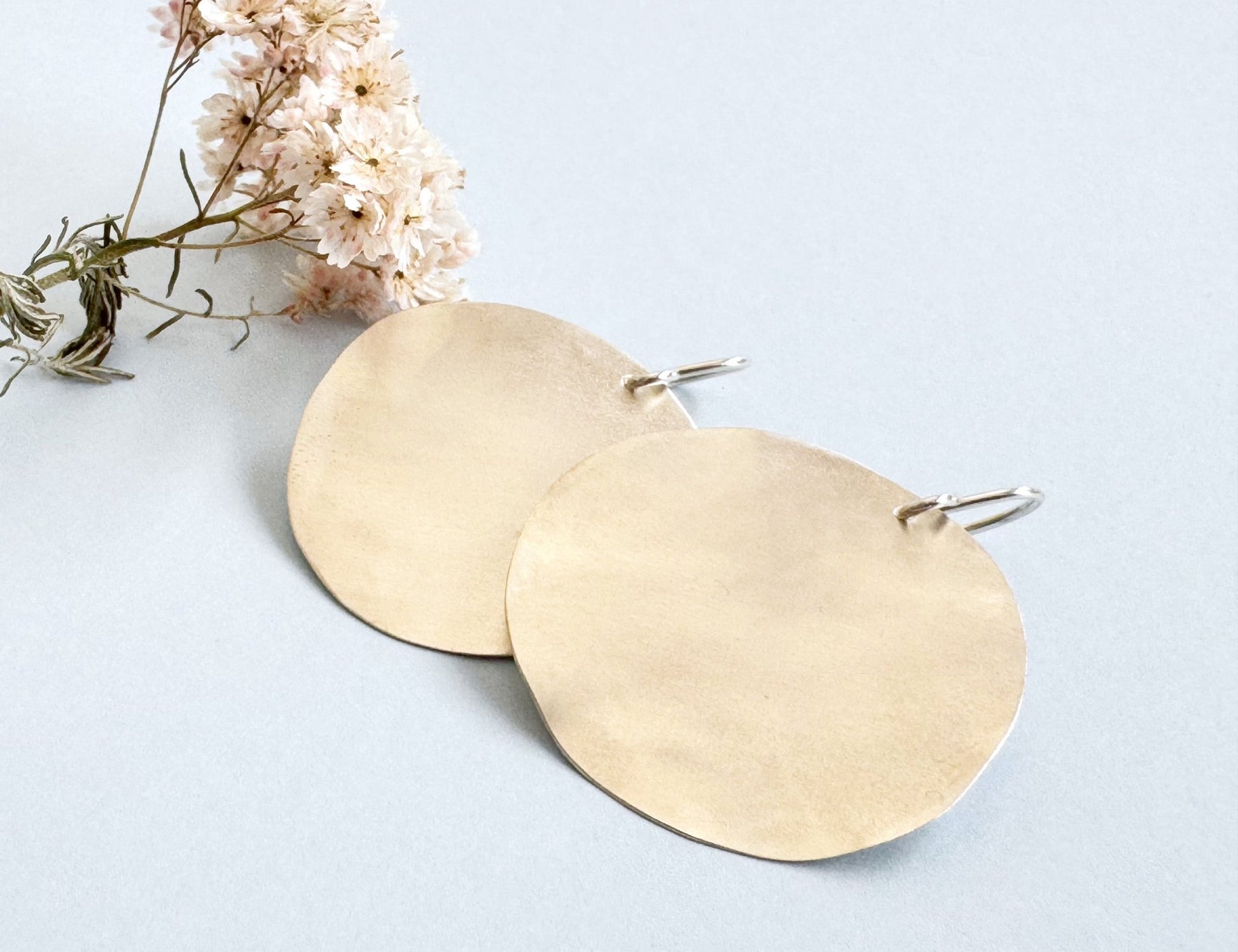 
A pair of large, hammered sterling silver disc earrings rests on a light blue background next to a small sprig of dried pink and white flowers.