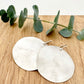 A pair of large, hammered, brush sterling silver disc earrings rests on a wooden surface next to a small sprig eucalyptus leaves.