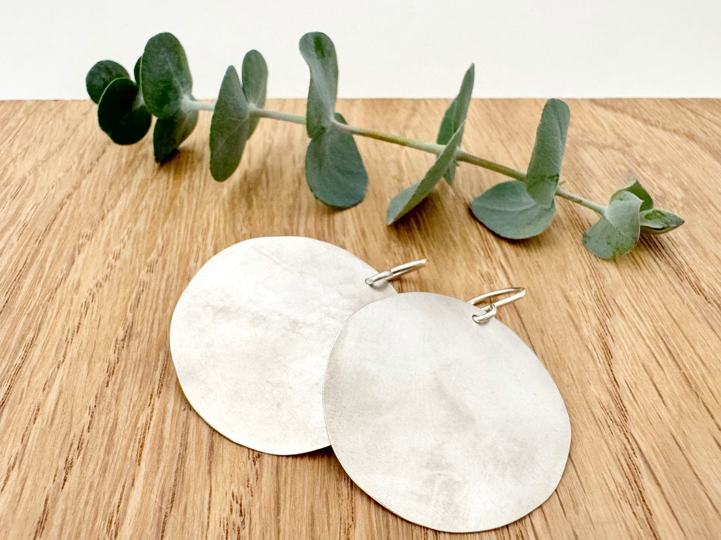 A pair of large, hammered, brush sterling silver disc earrings rests on a wooden surface next to a small sprig eucalyptus leaves.