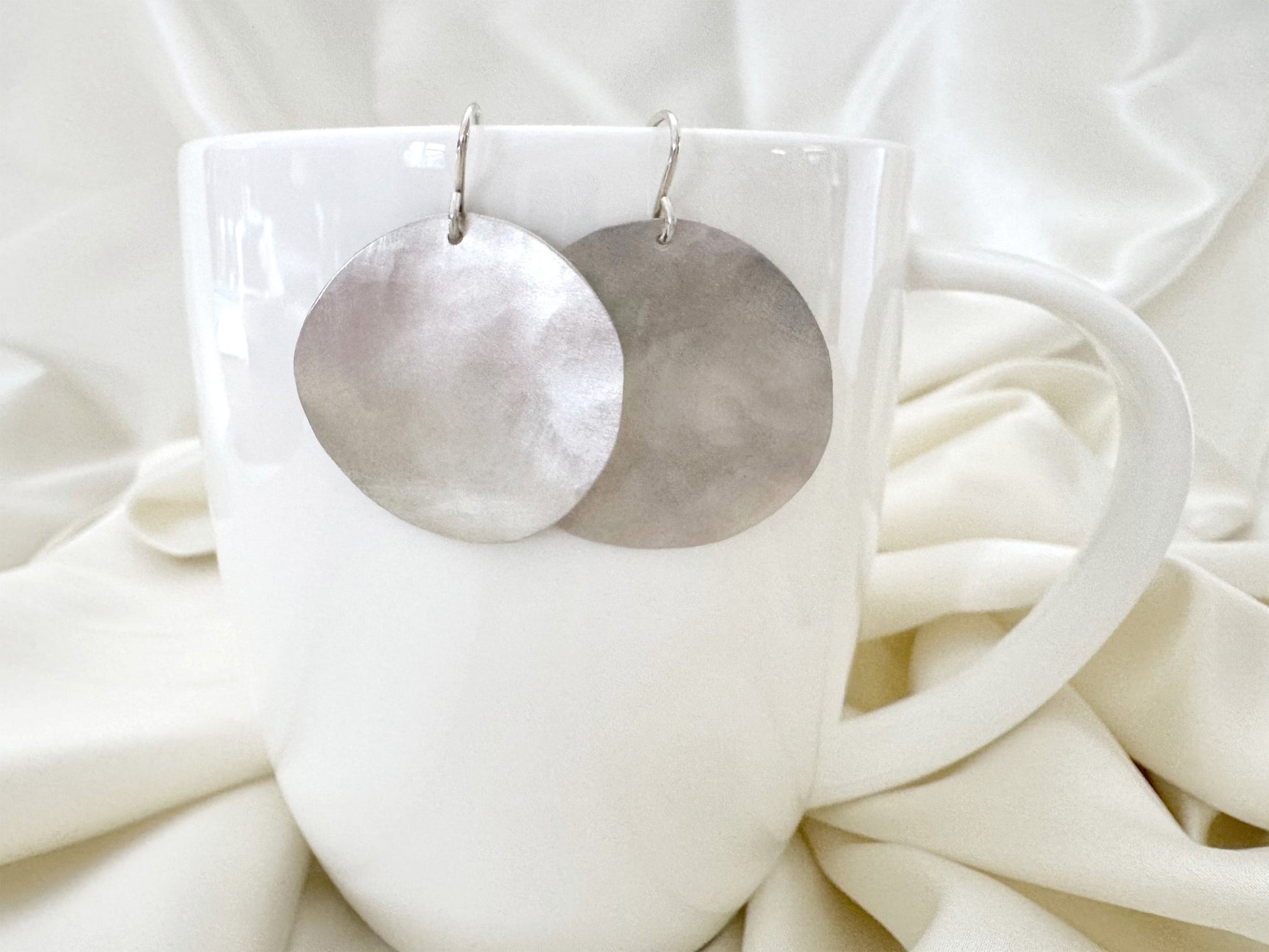Silver circular earrings on a white cup with a white fabric background