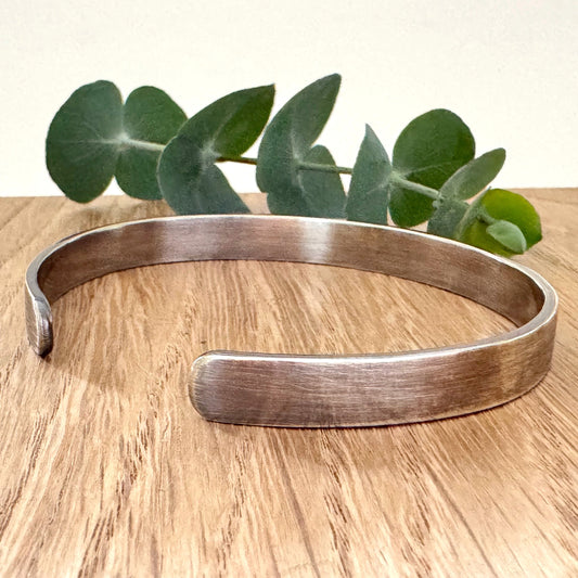 An oxidised sterling silver cuff bracelet on a wooden surface with green leaves in the background.