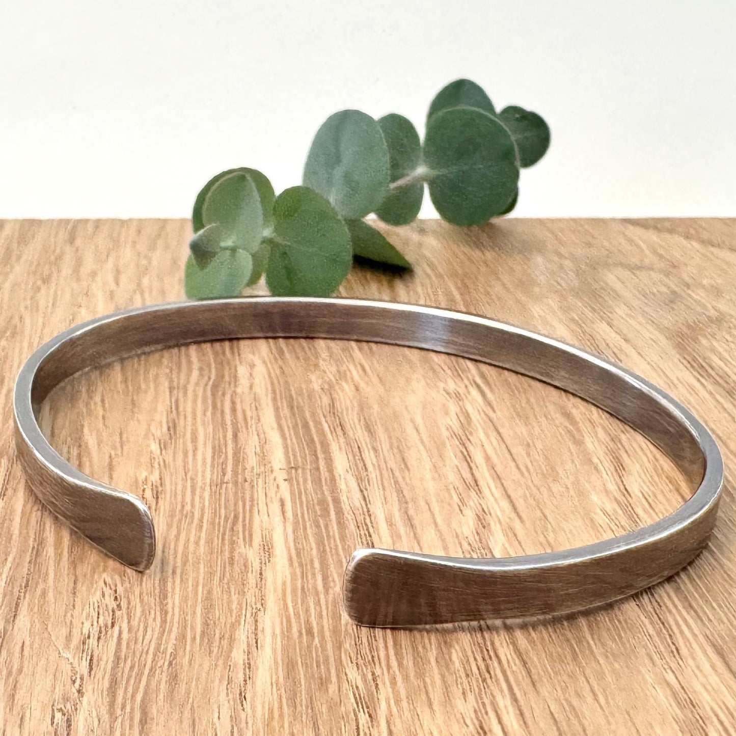 A sterling silver bracelet with flared ends and an oxidised finish, on a wooden surface with a small sprig eucalyptus leaves in the background.