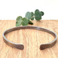 An oxidised sterling silver bracelet with flared ends on a wooden surface with eucalyptus leaves in the background.