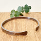 An oxidised sterling silver bracelet with flared ends on a wooden surface with eucalyptus leaves in the background.