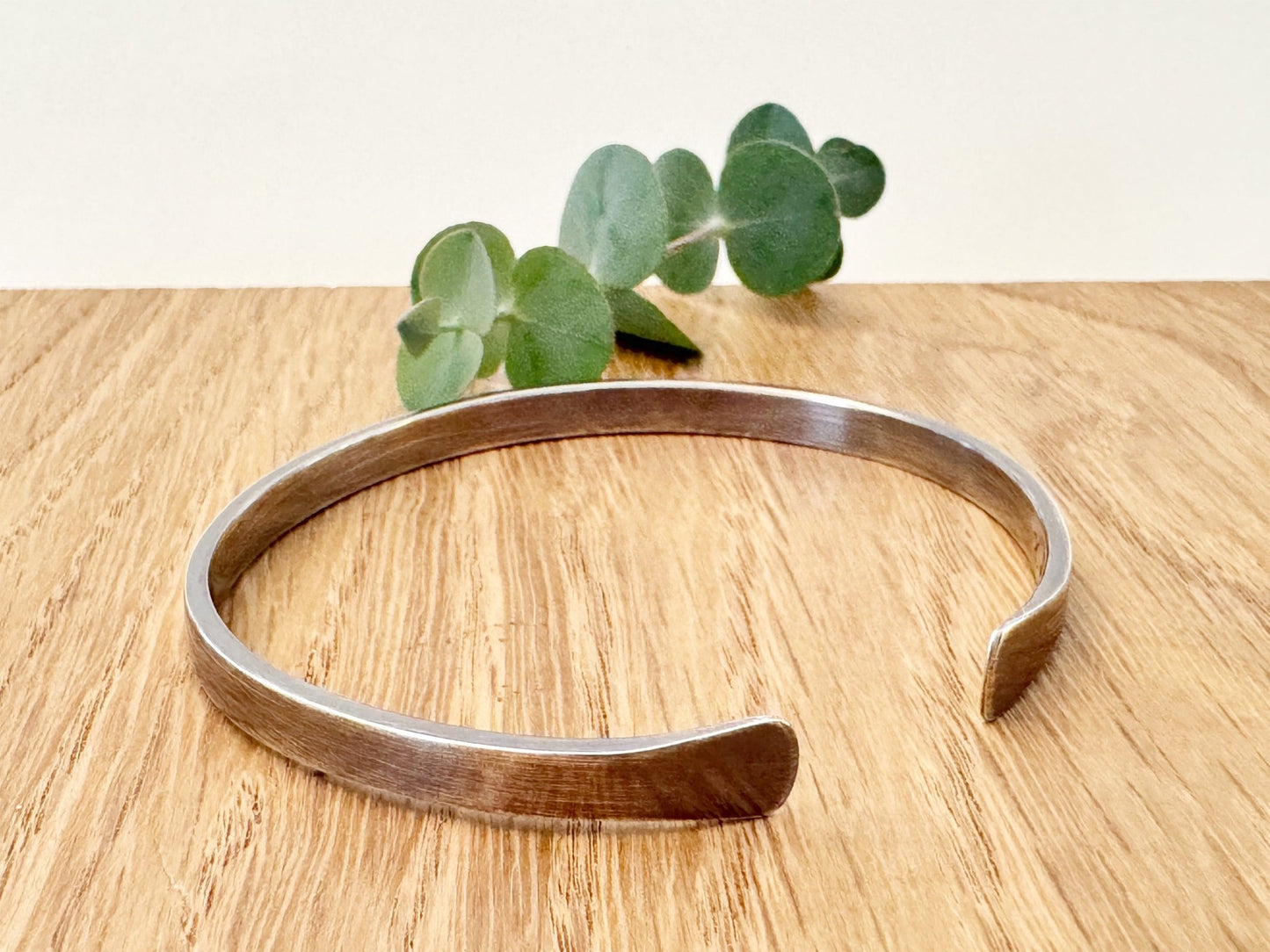 An oxidised sterling silver bracelet with flared ends on a wooden surface with eucalyptus leaves in the background.