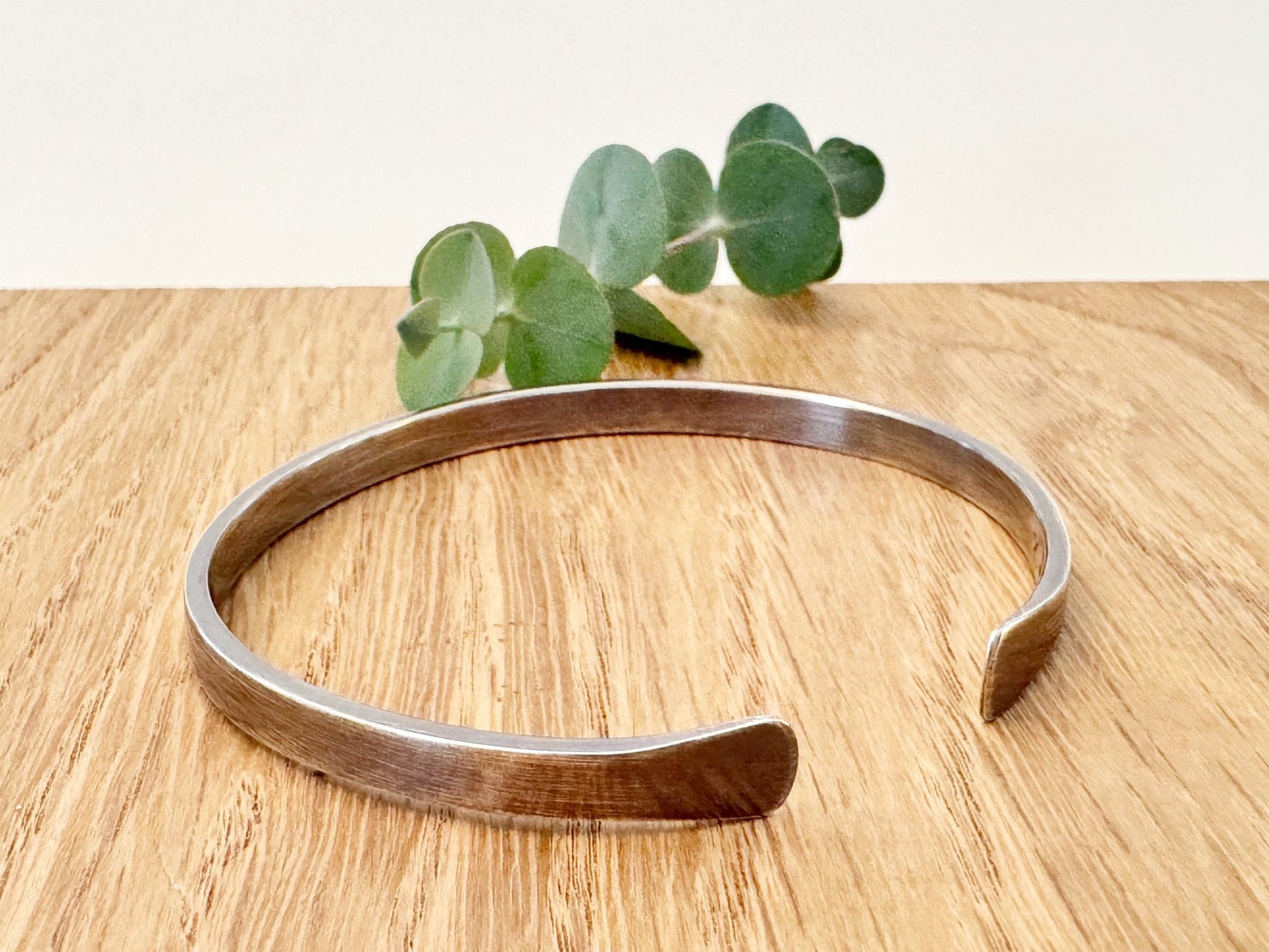 An oxidised sterling silver bracelet with flared ends on a wooden surface with eucalyptus leaves in the background.
