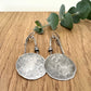 Pair of silver earrings on a wooden surface with a sprig of eucalyptus.