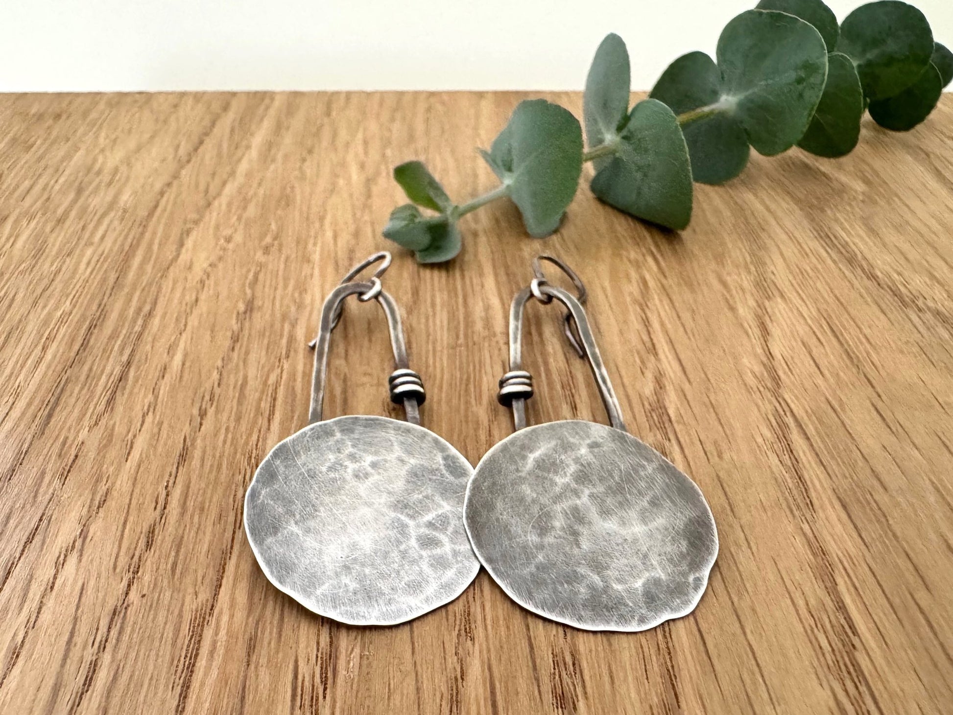 Pair of silver earrings on a wooden surface with a sprig of eucalyptus.