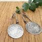 Pair of silver earrings on a wooden surface with eucalyptus leaves.