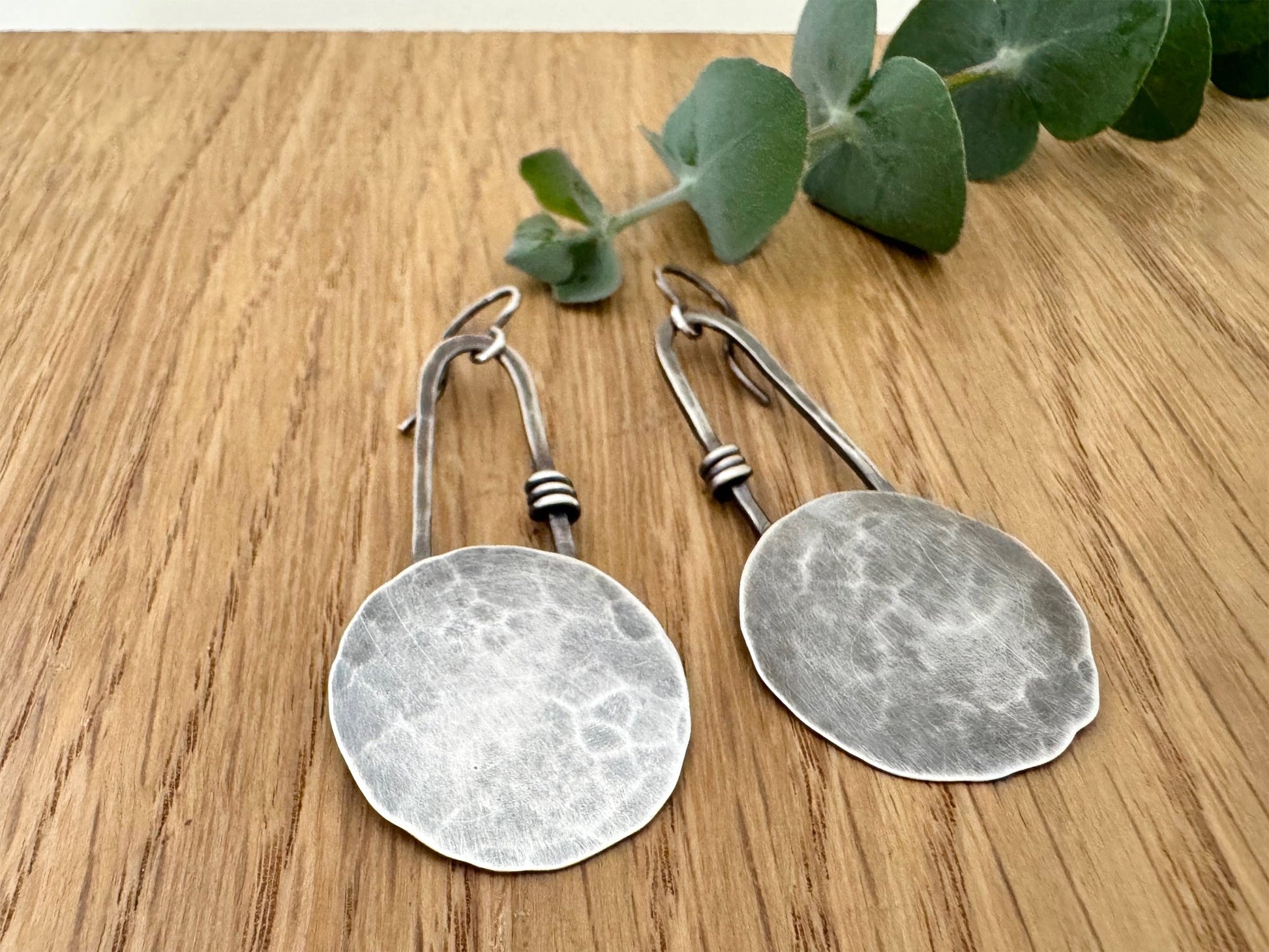 Pair of silver earrings on a wooden surface with eucalyptus leaves.