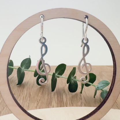 Sterling silver earrings with a treble clef design on a wooden surface with a small sprig of eucalyptus leaves.